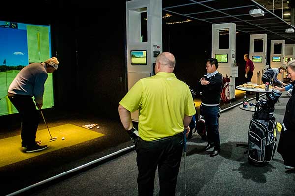 A group of people standing around watching a man prepare to swing at a golf ball indoors in front of a screen
